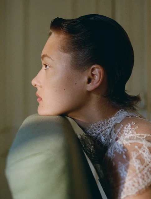 Elegant portrait of a woman with sleek dark hair, wearing a lace top, resting her head against a light green cushion in soft light.