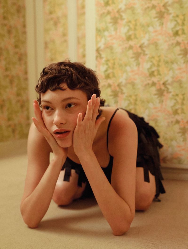 Young woman in black dress kneeling indoors, looking to the side with hands to face, floral wallpaper background in a medium shot.