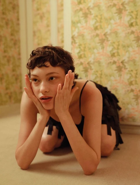 Young woman in black dress kneeling indoors, looking to the side with hands to face, floral wallpaper background in a medium shot.