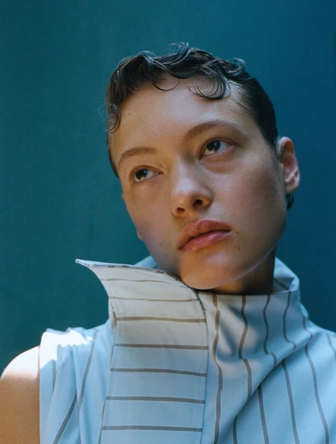 Close-up studio portrait of a young adult woman with dark, short, wet-look wavy hair, wearing a striped sleeveless top, against a teal background.