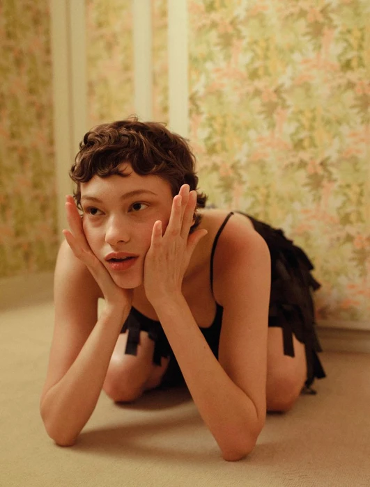 Model Daria Zolotova kneeling on a carpet with hands touching her face, wearing a black slip dress against floral wallpaper.