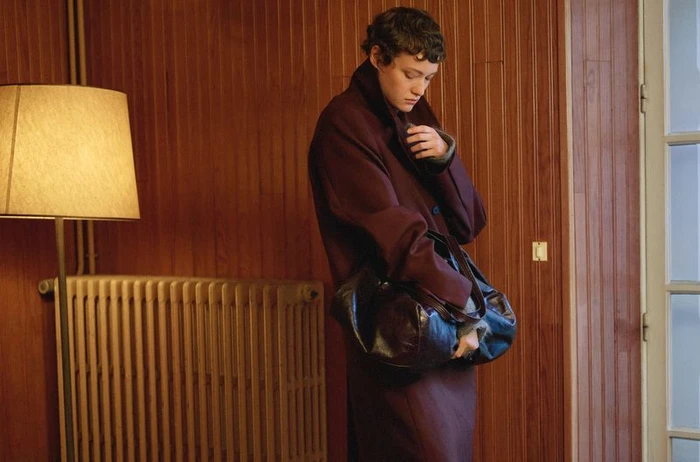 Model Daria Zolotova wearing an oversized burgundy coat and holding a dark leather bag in front of a wood-paneled wall.