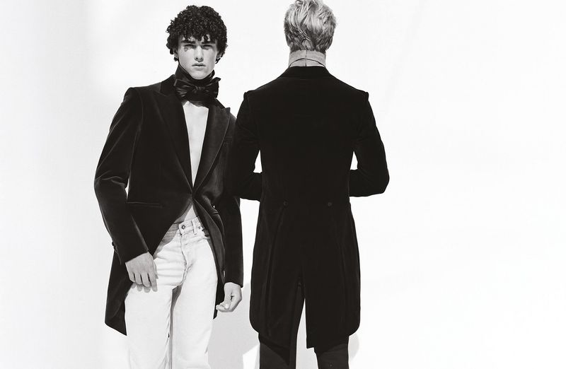 Two adult men in formal wear stand against a bright backdrop, one facing forward and one facing away, presenting a study in contrasts.