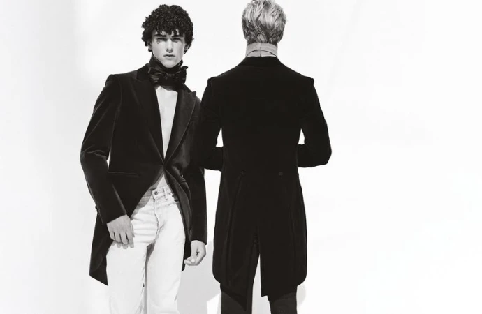Two adult men in formal wear stand against a bright backdrop, one facing forward and one facing away, presenting a study in contrasts.