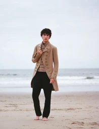 Model Xavier Karwas barefoot on a sandy beach wearing a tailored beige tailcoat and black trousers.