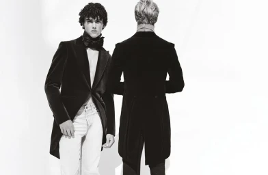 Two male models posing in black velvet tailcoats against a plain white studio backdrop for a fashion editorial.