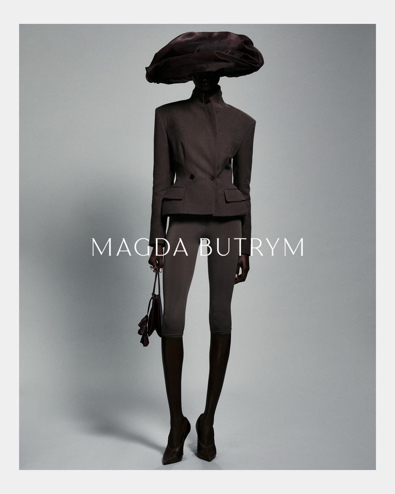A fashion shot showcases a model in a brown double-breasted blazer, matching leggings, heels, and a large hat, against a grey backdrop.