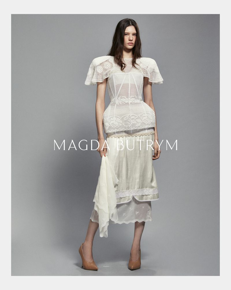 A woman wearing a white lace top and skirt stands in a photo studio with neutral background, showcasing a fashion editorial look.