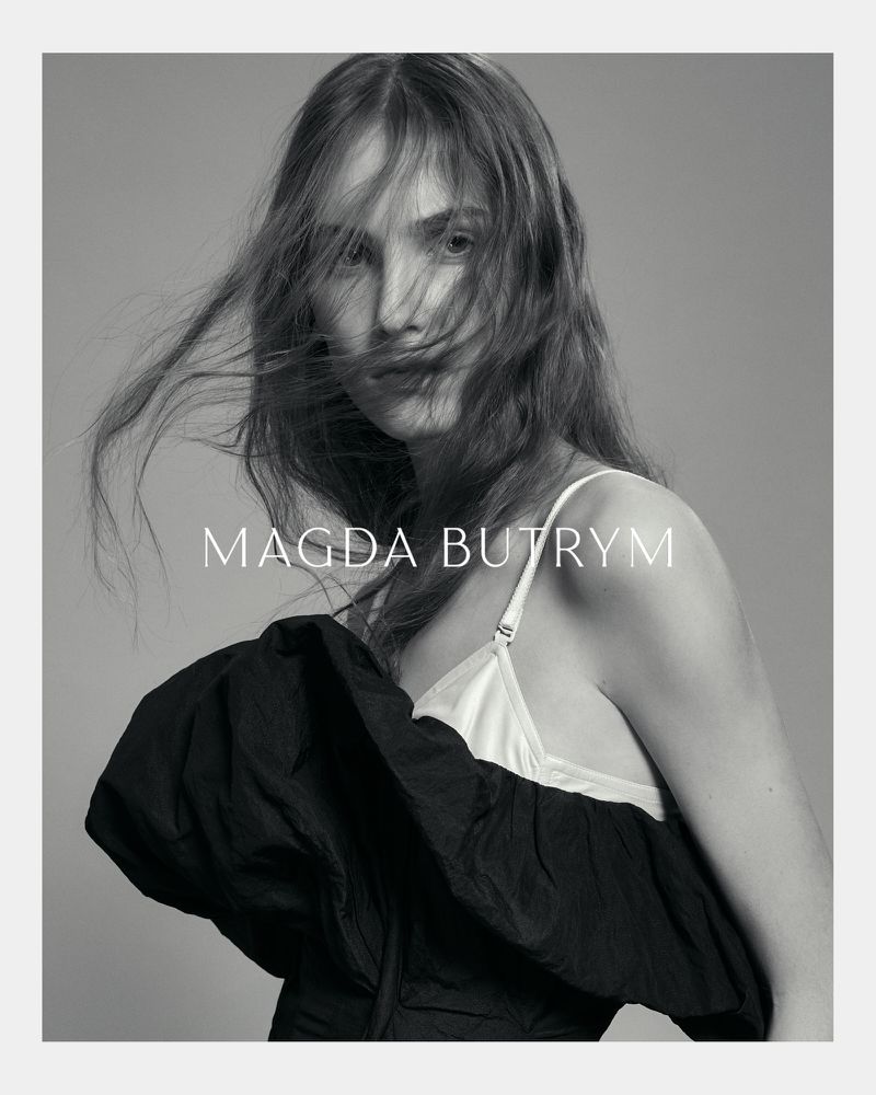 Portrait of a woman with long flowing hair wearing a black top with a white camisole, posing in a photo studio with a grey backdrop.
