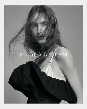 Model with long tousled hair wearing a white top and dark ruffled garment against a grey background with brand text
