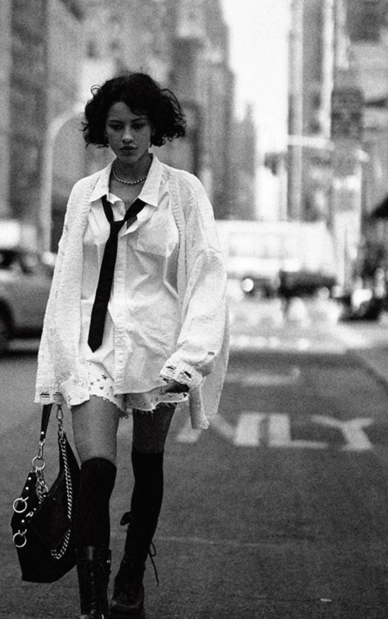 Woman in oversized white shirt and black tie walks city street with shoulder bag. Black and white fashion editorial photography.