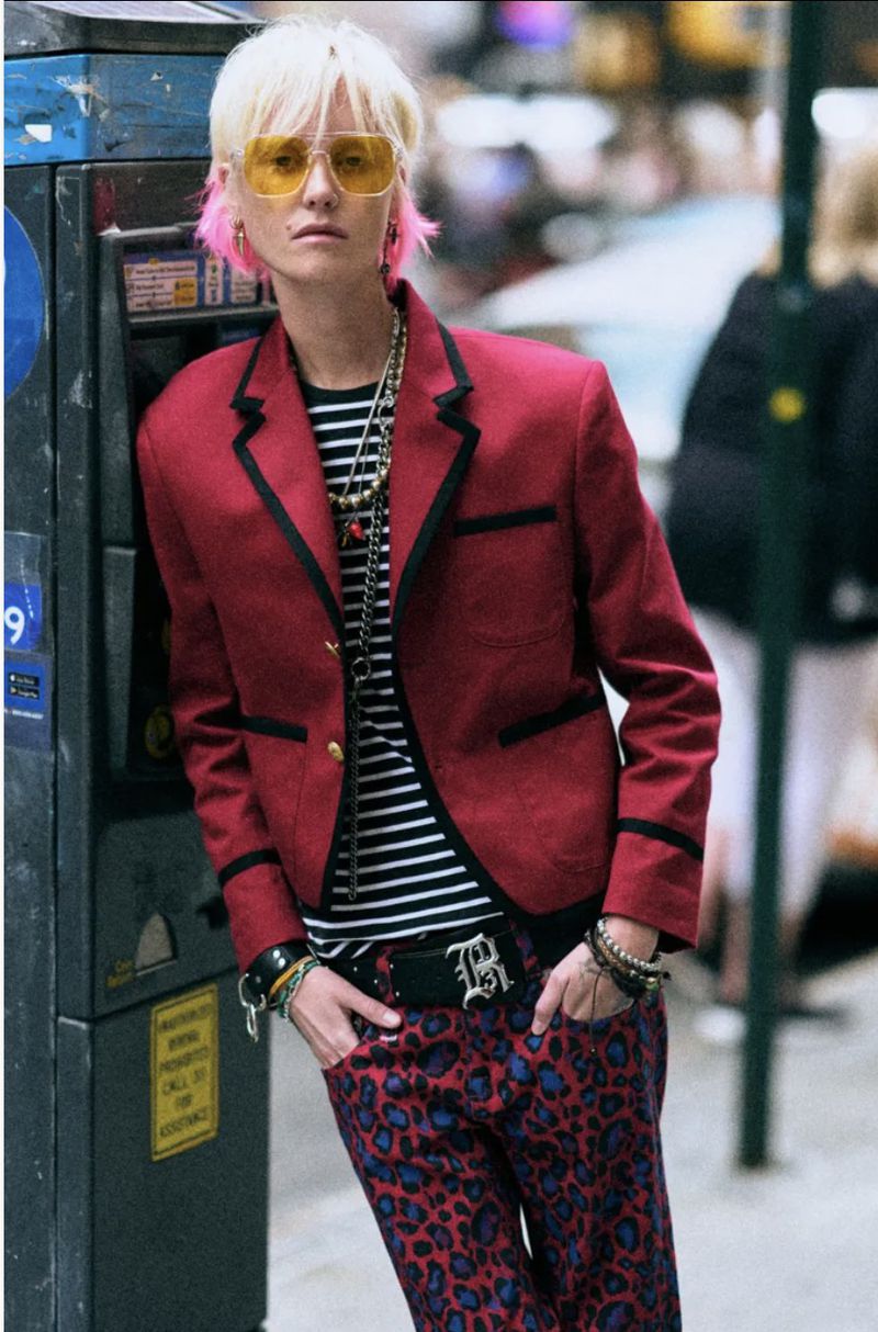 A stylish adult in a red blazer, black and white striped shirt, and patterned jeans leans against a coin-operated telephone in the city.