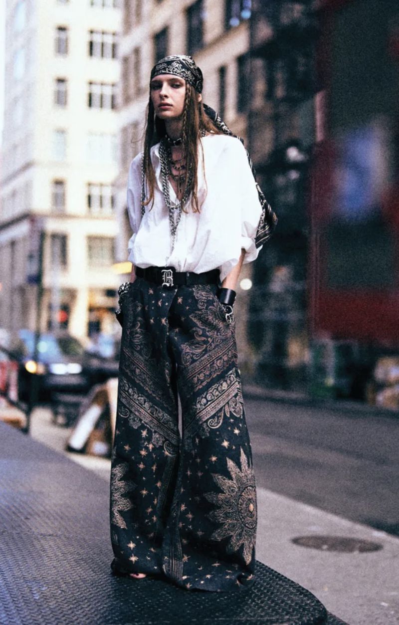 Model wearing a white button-up shirt, headscarf, and oversized patterned wide-leg trousers on a city street.