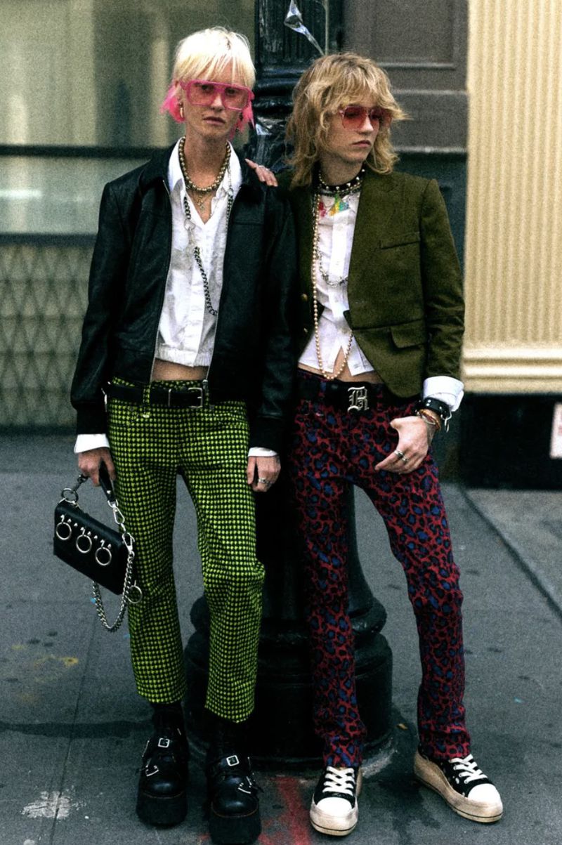 Two young adults in stylish outfits pose outdoors with one's arm around the other, wearing colorful pants and accessorizing with unique accessories.