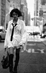 Model walking down a city street wearing an oversized white shirt, black tie, knee-high socks, and carrying a black handbag