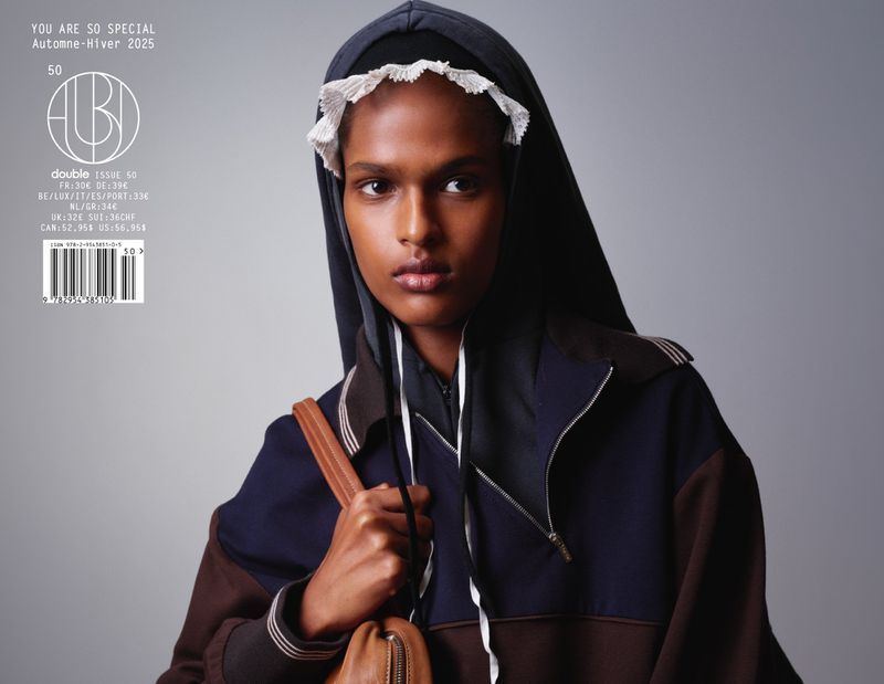Model wearing a dark hooded track jacket with white lace bonnet trim and a brown leather shoulder bag against a grey background.