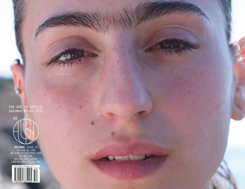 Close-up portrait of a young adult female face with pierced ears, mole on cheek, light skin, light pink lips with magazine cover text.
