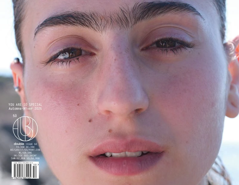 Close-up portrait of a young adult female face with pierced ears, mole on cheek, light skin, light pink lips with magazine cover text.