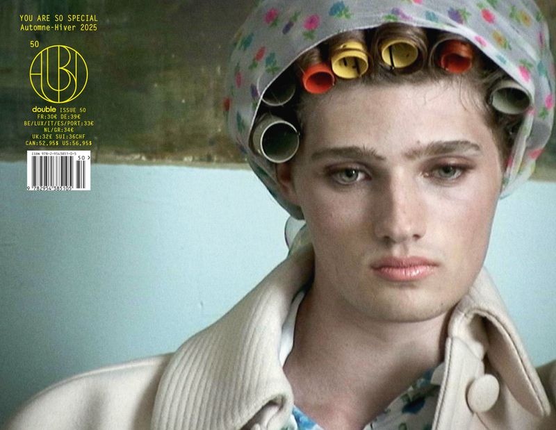 Close-up studio shot of a model wearing a floral headscarf with hair curlers, a patterned top, and a white coat against a pale blue backdrop.