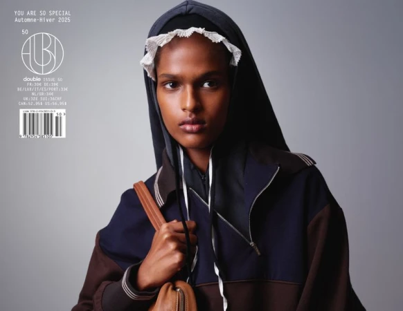 Model wearing a dark hooded track jacket with white lace bonnet trim and a brown leather shoulder bag against a grey background.
