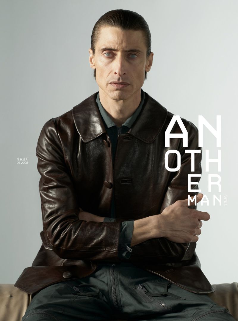 Portrait of a man with slicked-back dark hair wearing a dark brown leather jacket against a light grey background.