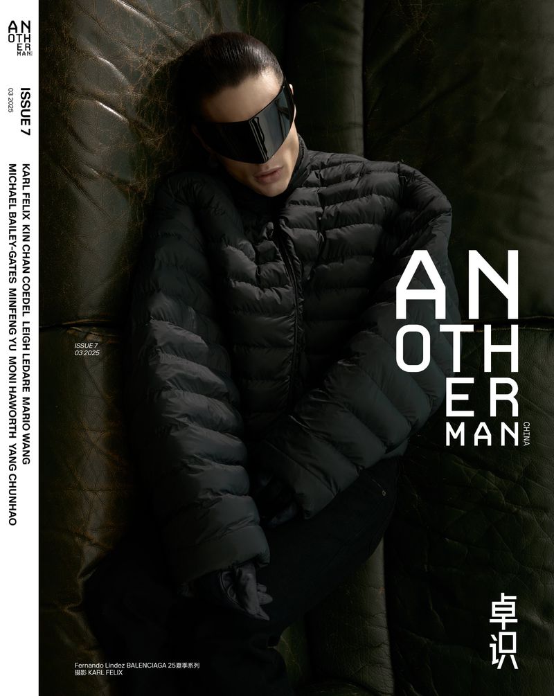 Magazine cover featuring a human wearing a black puffer jacket and futuristic visor, lying on a textured surface. Dark, editorial fashion.