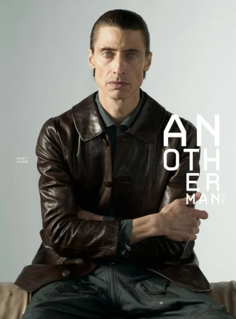 Portrait of a man with slicked-back dark hair wearing a dark brown leather jacket against a light grey background.