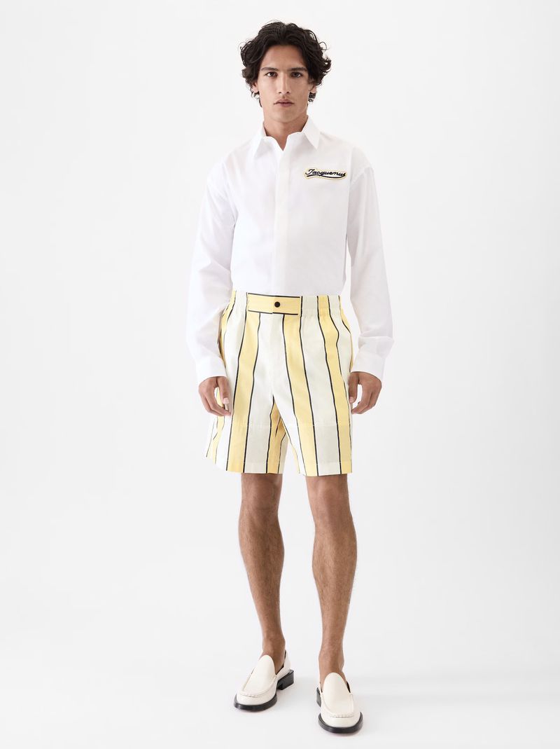 Full shot of a young adult male model in a white button-down shirt and striped shorts, posing in a bright white photo studio.