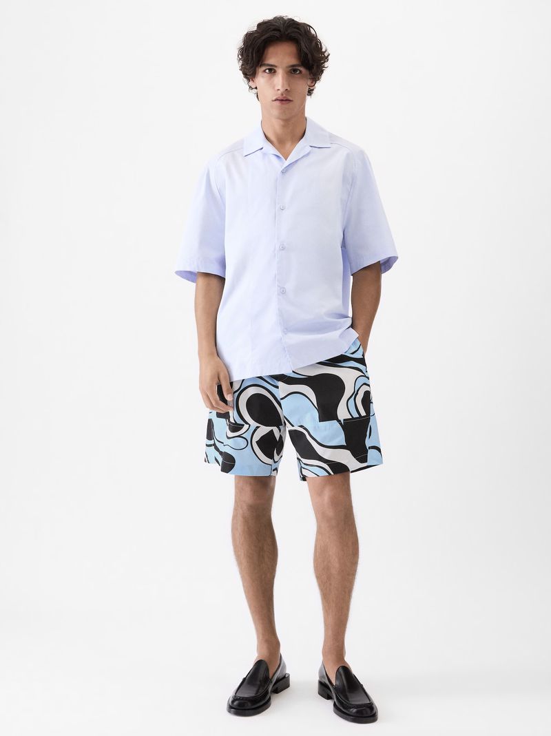Model Morgan Sahadow in a light blue short-sleeved button-up shirt and patterned shorts against a white background.