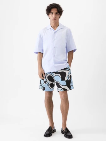 Model Morgan Sahadow in a light blue short-sleeved button-up shirt and patterned shorts against a white background.