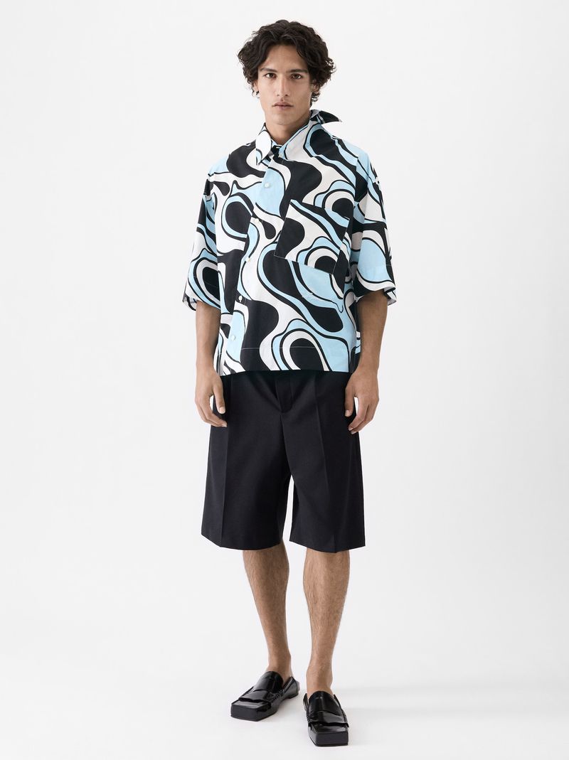 A young adult male stands in a full shot against a white backdrop, wearing a blue, black, and white patterned shirt and black shorts.