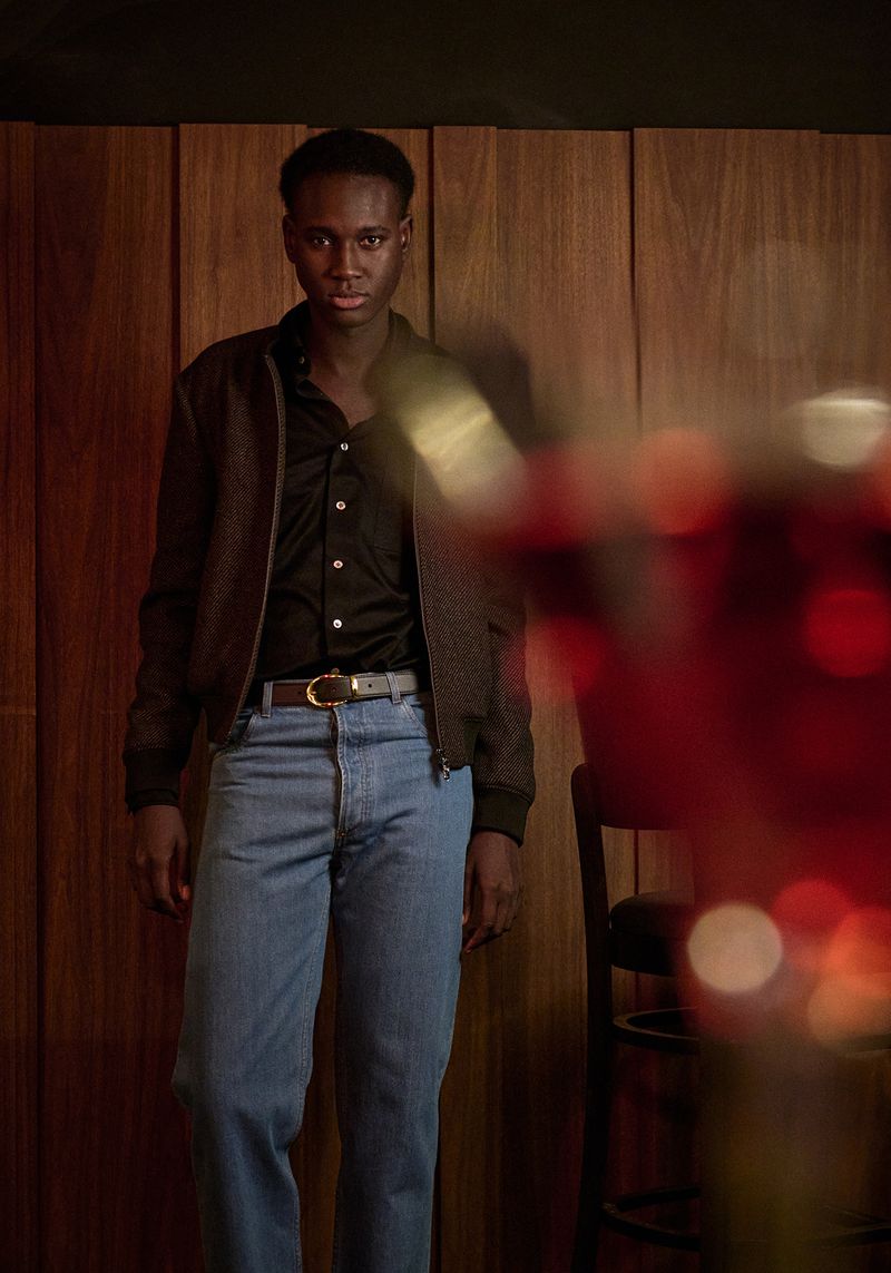 Portrait of a Black man in a brown jacket and blue jeans, standing indoors against a wood-paneled wall, with a blurred drink in the foreground.