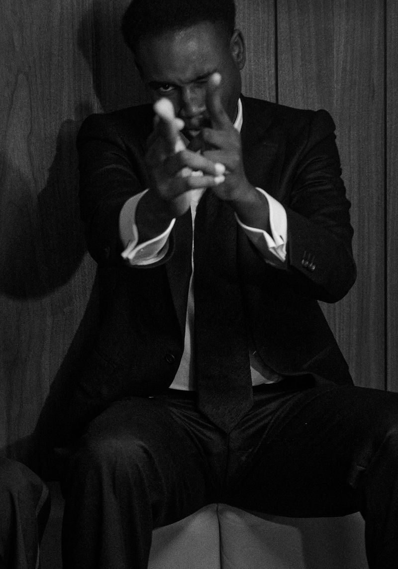 Black and white portrait of an adult male in a suit aiming finger guns indoors against a wood-paneled wall, creating a dramatic effect.