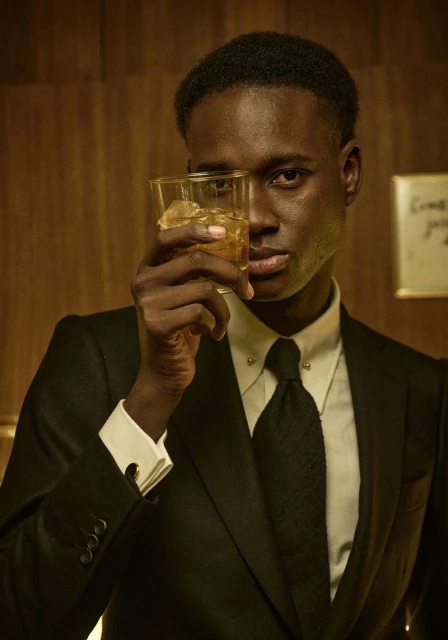 Stylish man in a suit holds a glass of amber liquor with ice, standing indoors with wood-panelled walls and soft, warm lighting.
