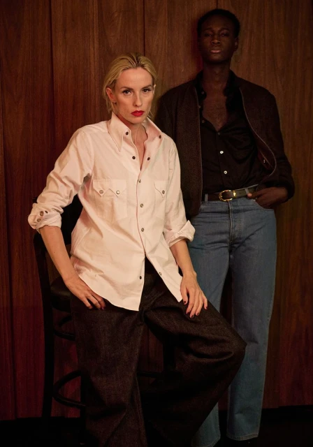 Two fashion models in a studio, one blonde female seated, one dark-skinned male standing against a wooden wall backdrop.