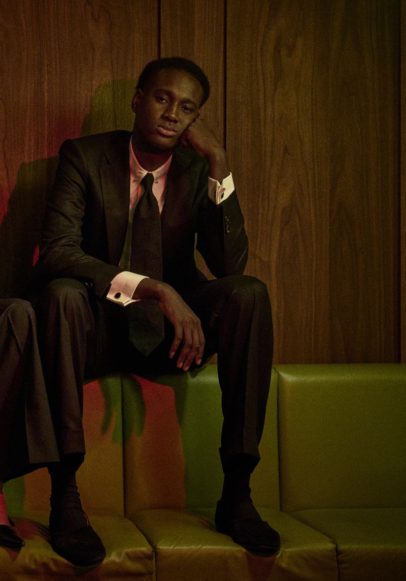 Portrait of a Black man in a suit sitting on a green banquette, with a wood paneled wall in the background and dramatic lighting.