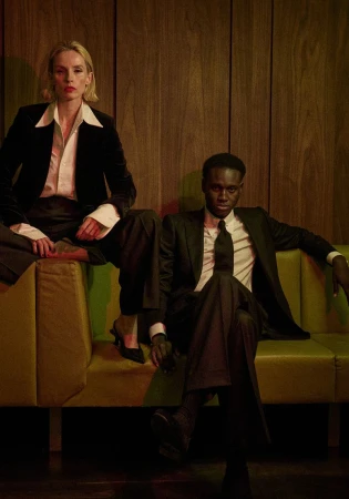 Two models in tailored black suits, including Aliou Gueye, seated on a green couch in front of a wood-paneled wall.