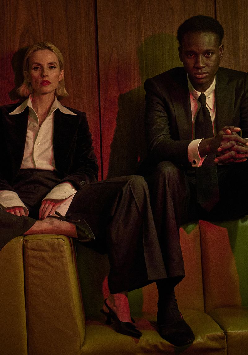 A black man and a white woman in suits sit on a retro couch in front of wood paneling in a fashion or celebrity and entertainment context.