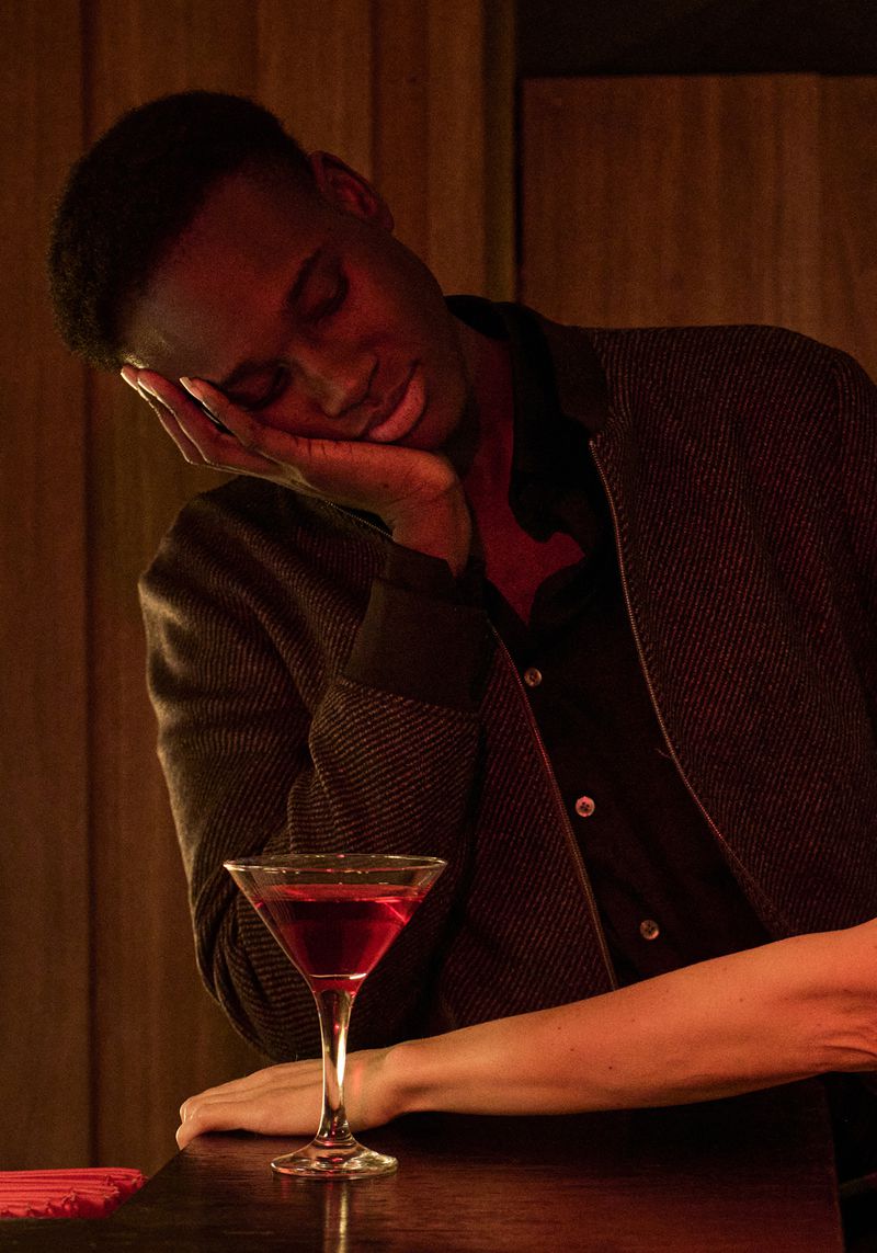 A young Black man rests his head on his hand next to a martini glass on a dimly lit bar, creating a mood of exhaustion or contemplation.