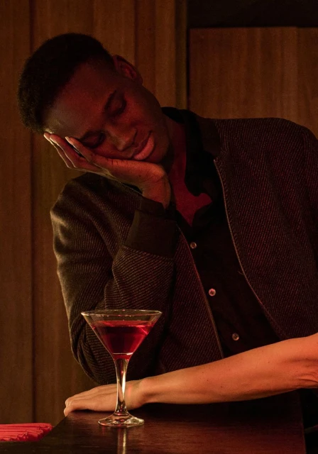 A young Black man rests his head on his hand next to a martini glass on a dimly lit bar, creating a mood of exhaustion or contemplation.