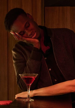 A young Black man rests his head on his hand next to a martini glass on a dimly lit bar, creating a mood of exhaustion or contemplation.