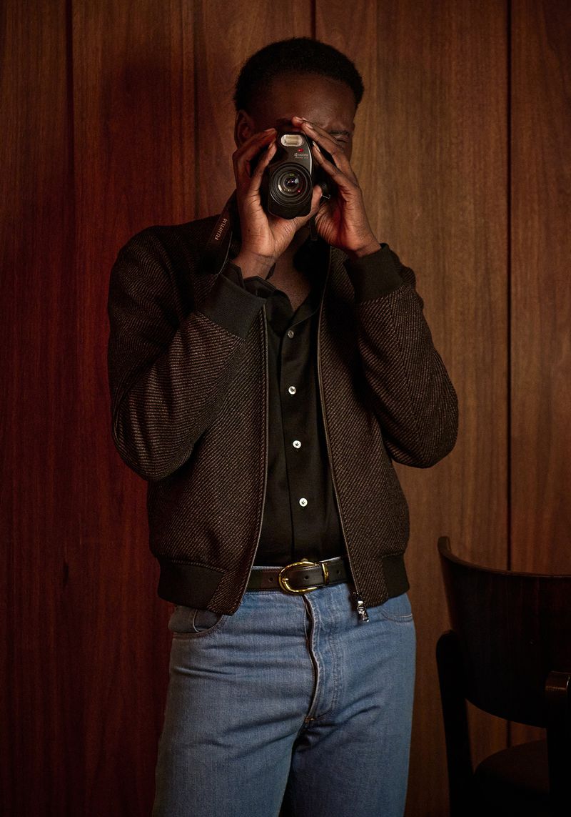 Dark-skinned adult male taking a picture with a compact camera indoors, wearing a jacket, blue jeans and a black button-down shirt.