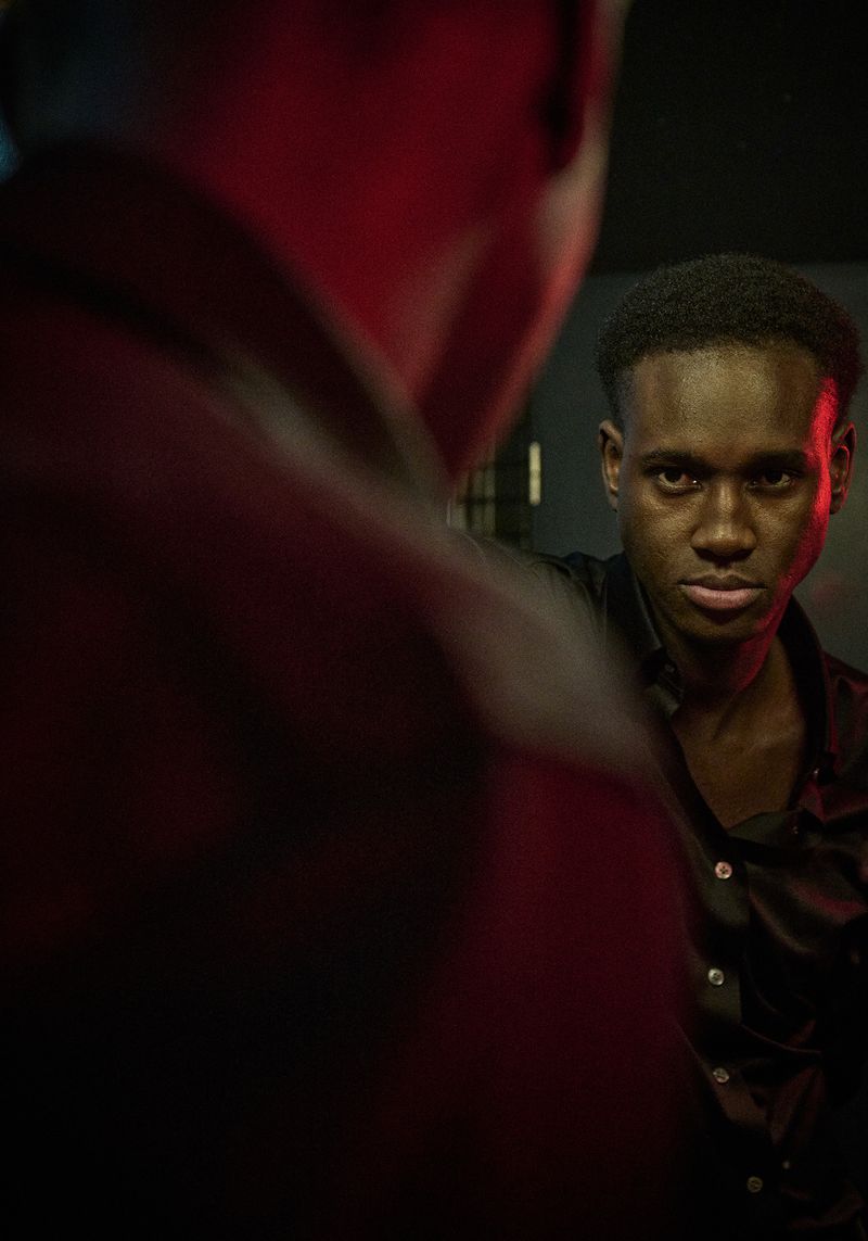 Portrait of a young Black man looking at his reflection in a mirror with a serious expression in a dark, indoor setting.