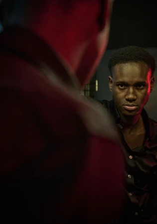 Portrait of a young Black man looking at his reflection in a mirror with a serious expression in a dark, indoor setting.