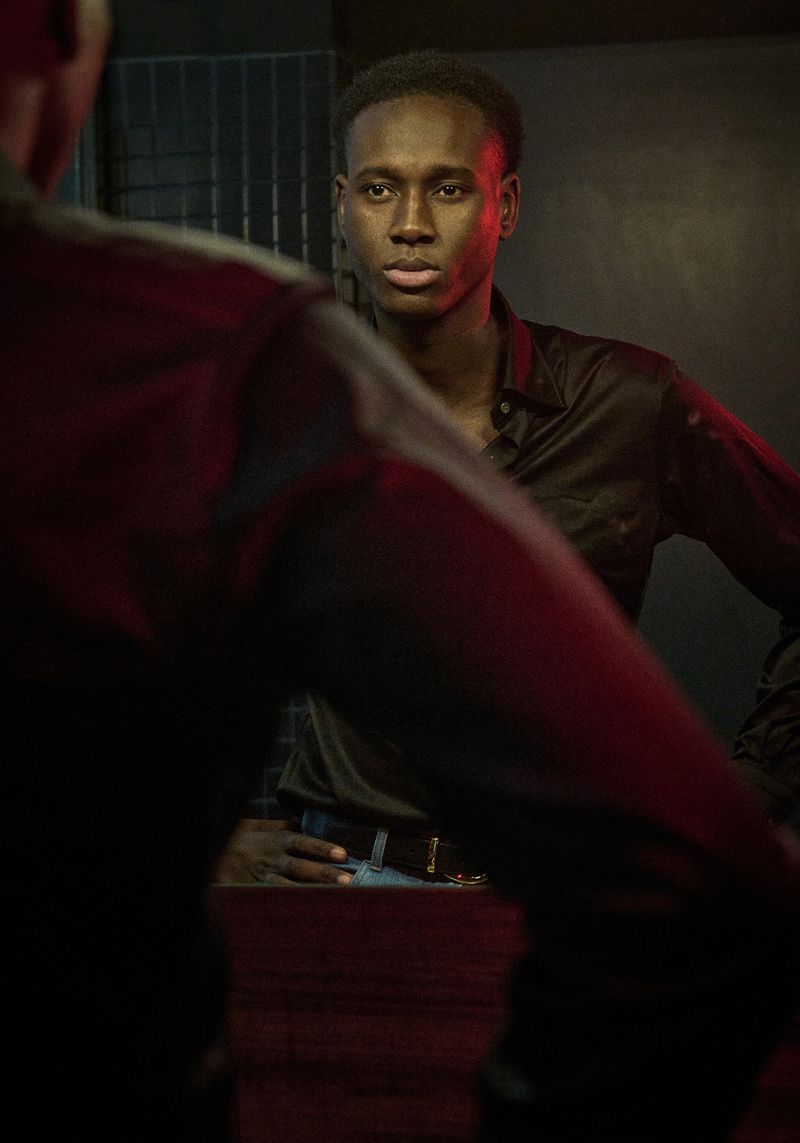 A dark-skinned male poses indoors, wearing a black button-down shirt, looking at his reflection in a mirror with red lighting in the background.