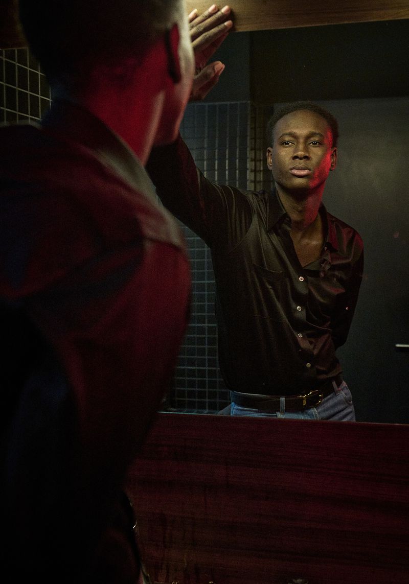 A young Black man in a satin shirt looks at his reflection in a mirror in a dimly lit interior setting with dark tiles.