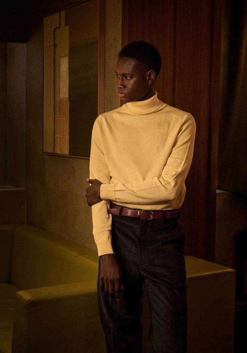 Young Black man poses indoors, wearing a yellow turtleneck sweater, black pants, and a brown belt, near yellow couch and patterned wall decor.