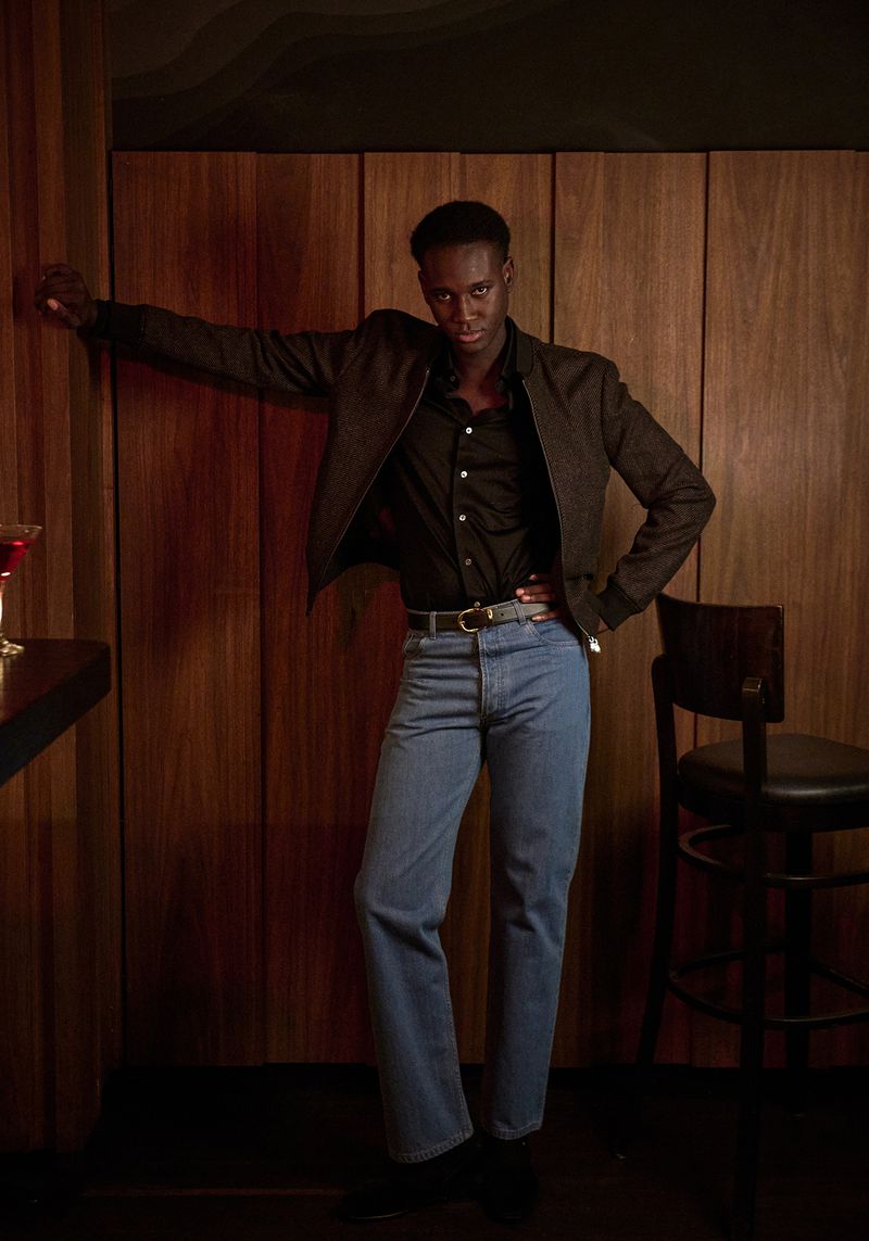 A stylish dark-skinned man in a jacket and jeans leans against a wood-paneled wall next to a bar table indoors, exuding a cool, casual vibe.