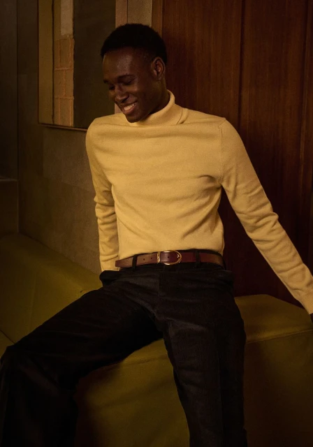 Stylish young Black man in a yellow turtleneck sweater and black trousers smiling while seated indoors, capturing a relaxed and joyful mood.