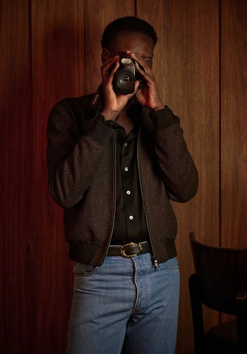 Model Aliou Gueye holding a vintage camera up to his face wearing a brown bomber jacket and blue jeans against a wood paneled wall.